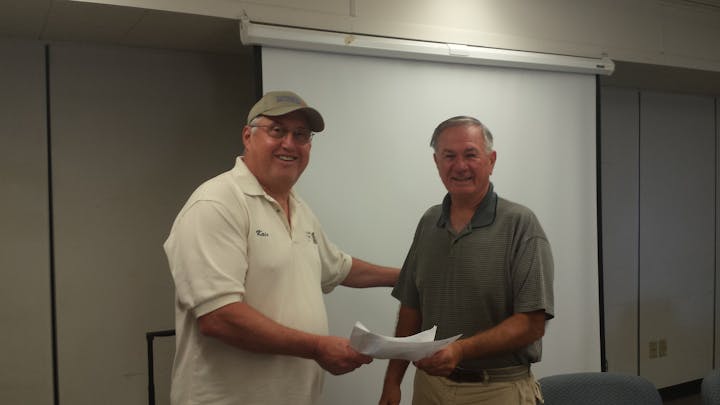 Two men shaking hands and exchanging papers in a room with a whiteboard.