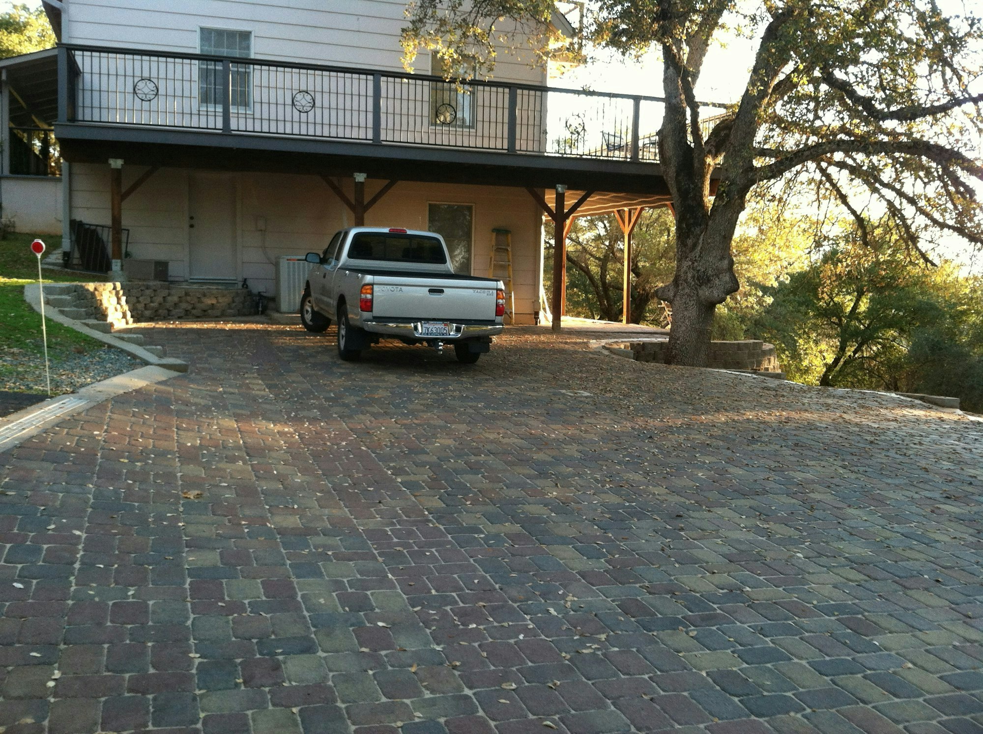 A pickup truck parked on a brick driveway in front of a house with a balcony and a large tree.