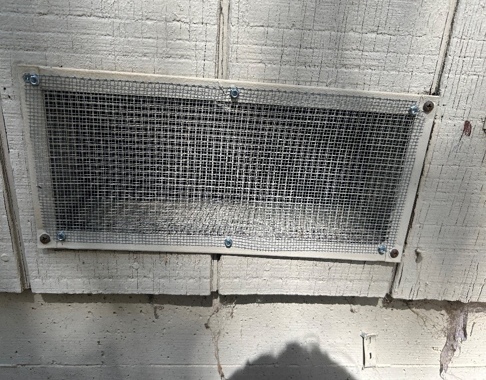 A metal mesh vent is attached to a wooden surface with screws.