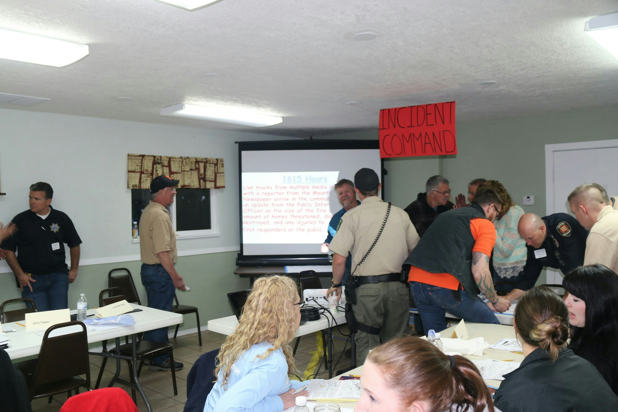 A group of people in a room with 'INCIDENT COMMAND' sign, looking at a presentation slide about a reported incident.