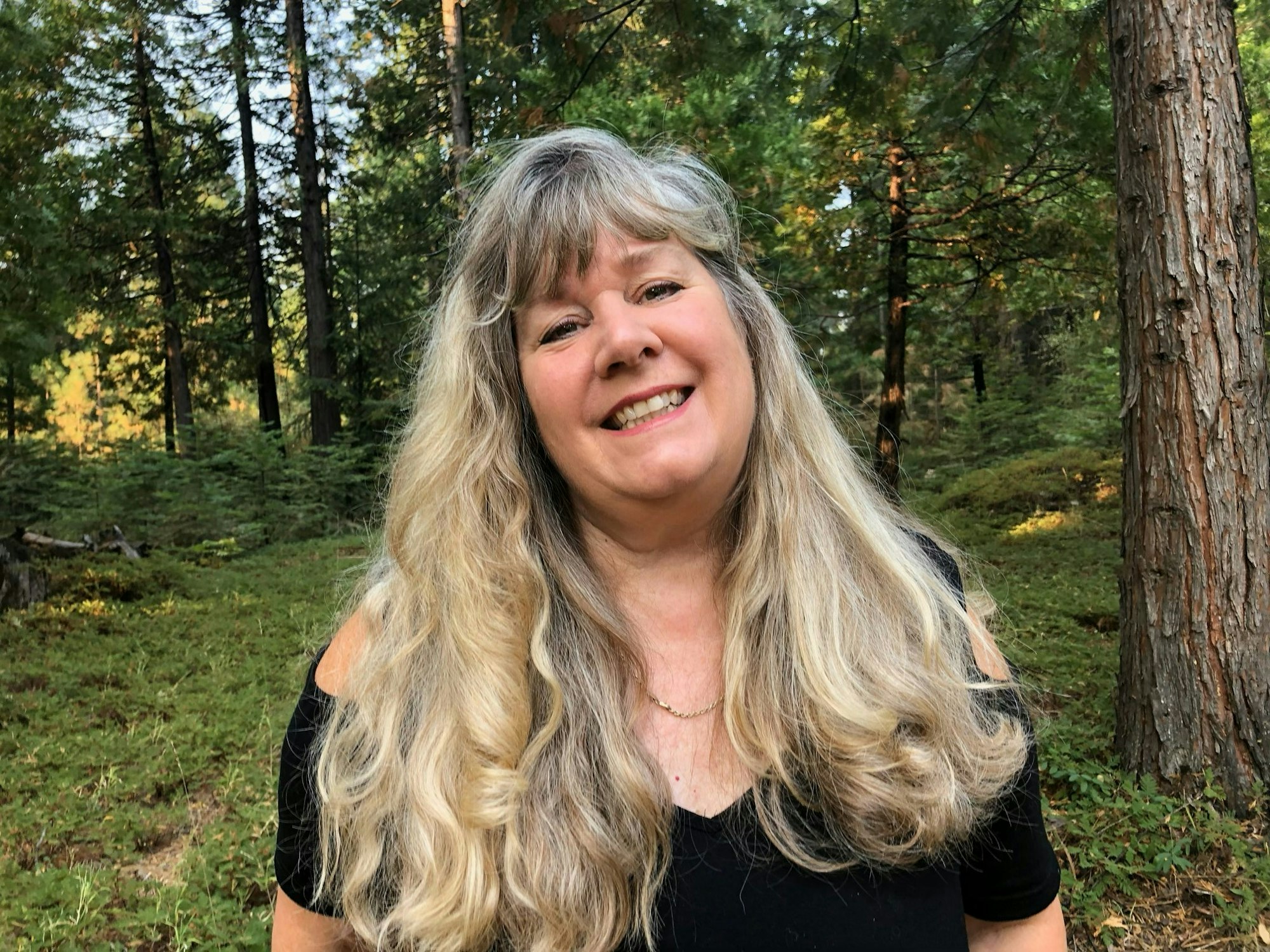 A smiling person with long, wavy hair standing in a forest with pine trees.