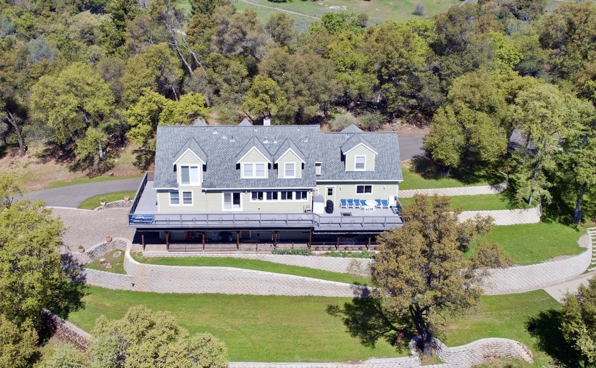 Aerial view of a large house with a blue-trimmed deck, surrounded by greenery and a cobblestone driveway.