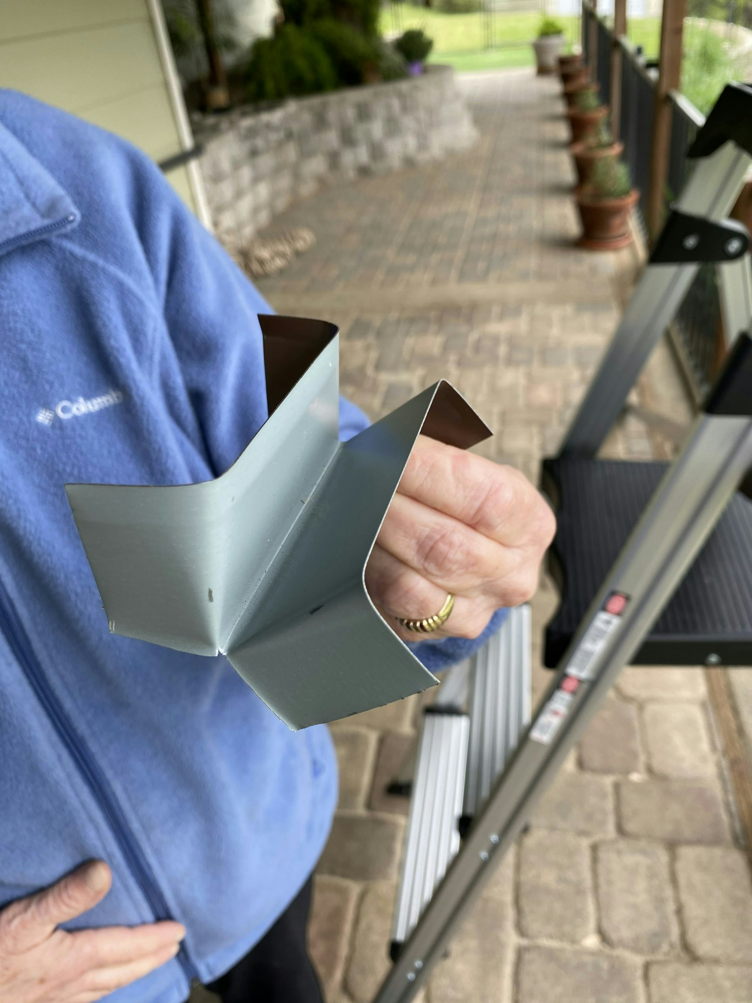 A person holding a metal channel or bracket, with a porch and potted plants in the background.