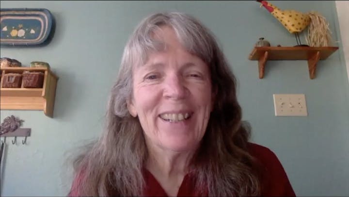 A smiling person with long hair in a room with decor: a shelf, a rooster ornament, and a clock.