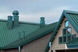 A building with green metal roofs, dormer windows, and vent pipes against a cloudy sky.