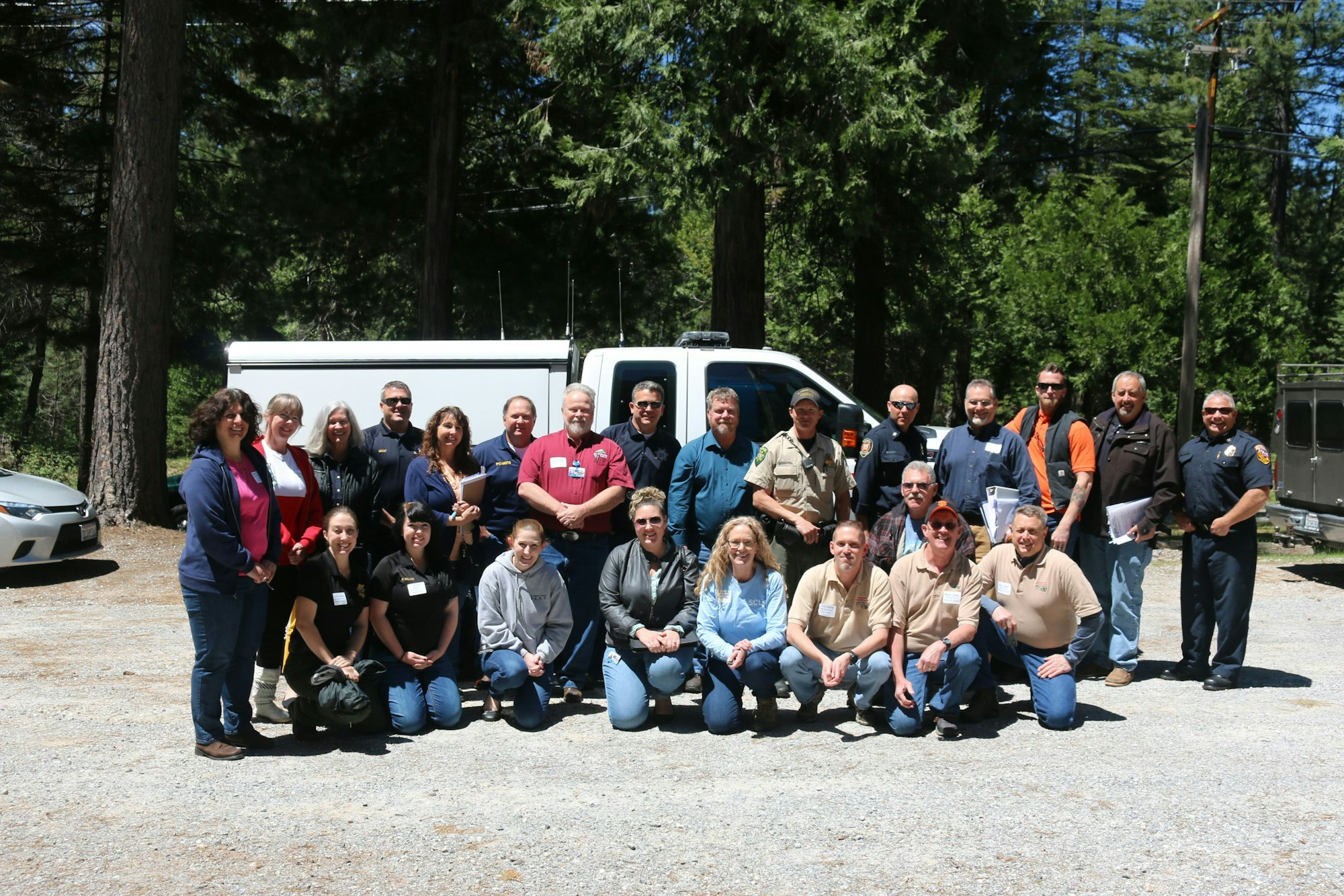 A group of people posing for a photo outdoors, with trees in the background and vehicles to the side.