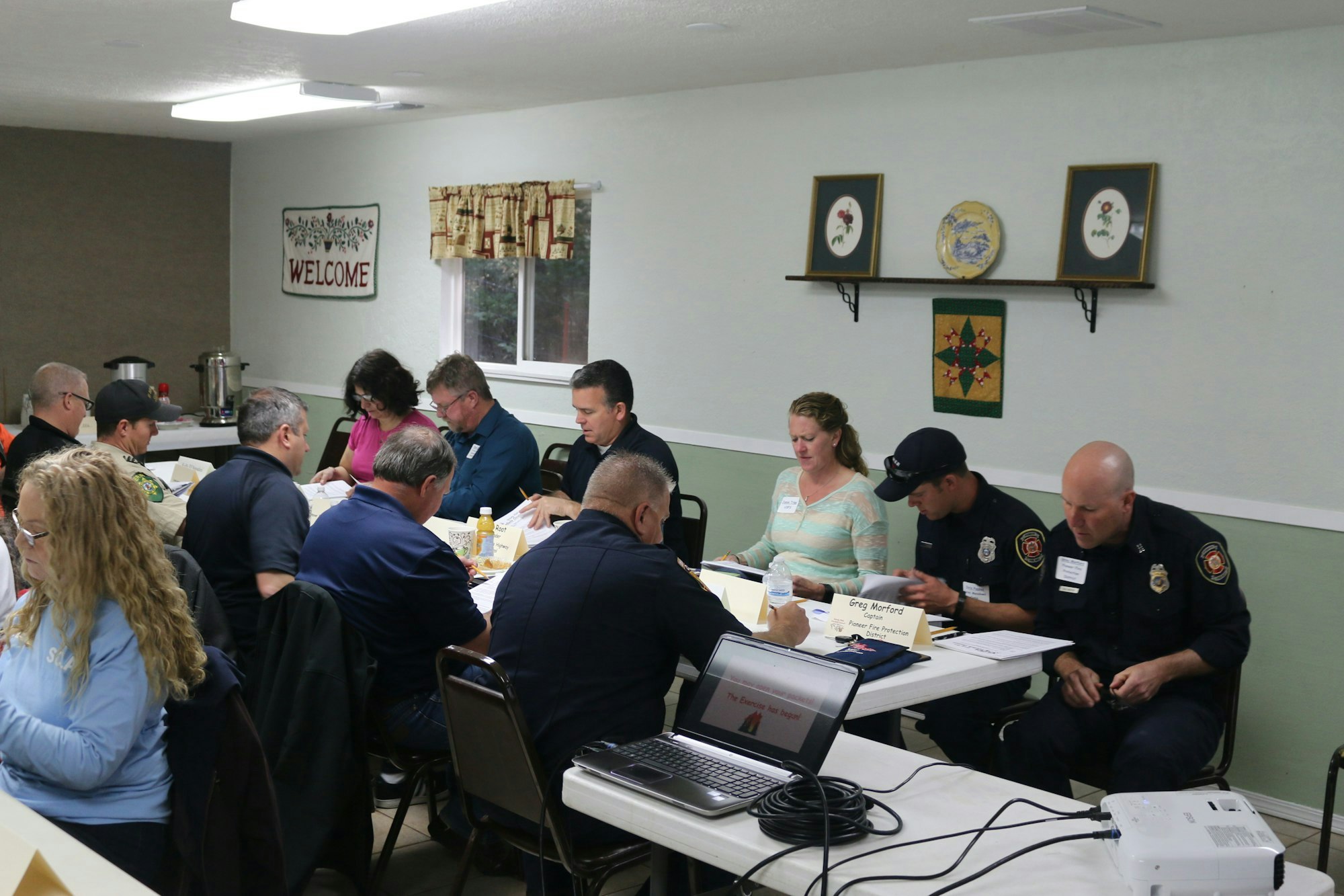 A group of people, including uniformed firefighters, seated at tables, focused on papers and discussions in a meeting room with a "WELCOME" sign.