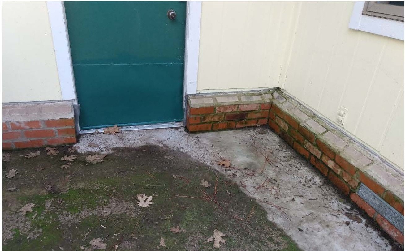 A green door with a brick border, concrete ground, and fallen leaves scattered around.