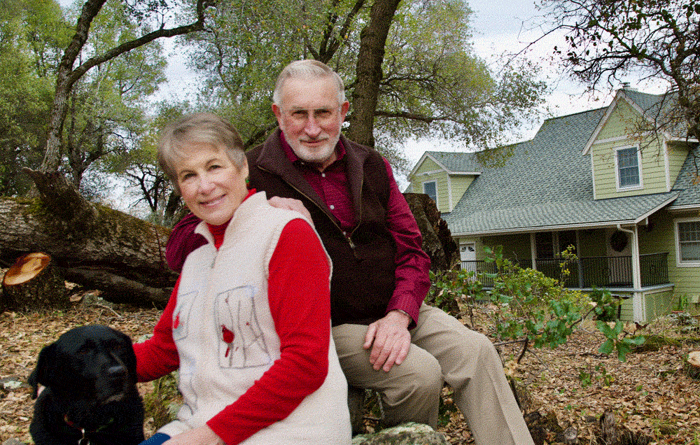 A couple with their dog in front of a house, surrounded by nature.