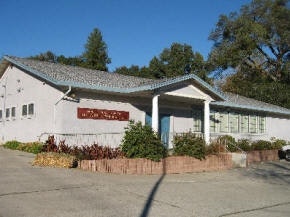 Single-story building with tiled roof, front porch, and landscaped garden under a blue sky.