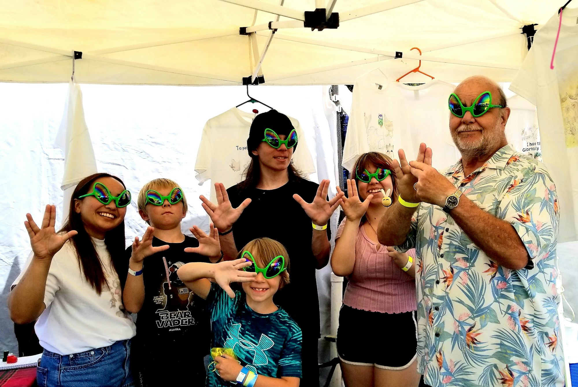 Group of six people wearing funky alien glasses, posing with hands up in a tent with t-shirts displayed.
