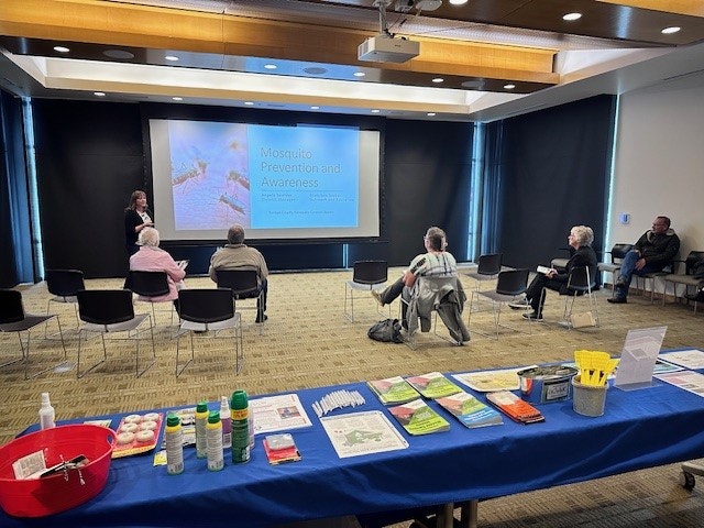 A presentation on Mosquito Prevention and Awareness with an audience and informational materials on a table in the foreground.