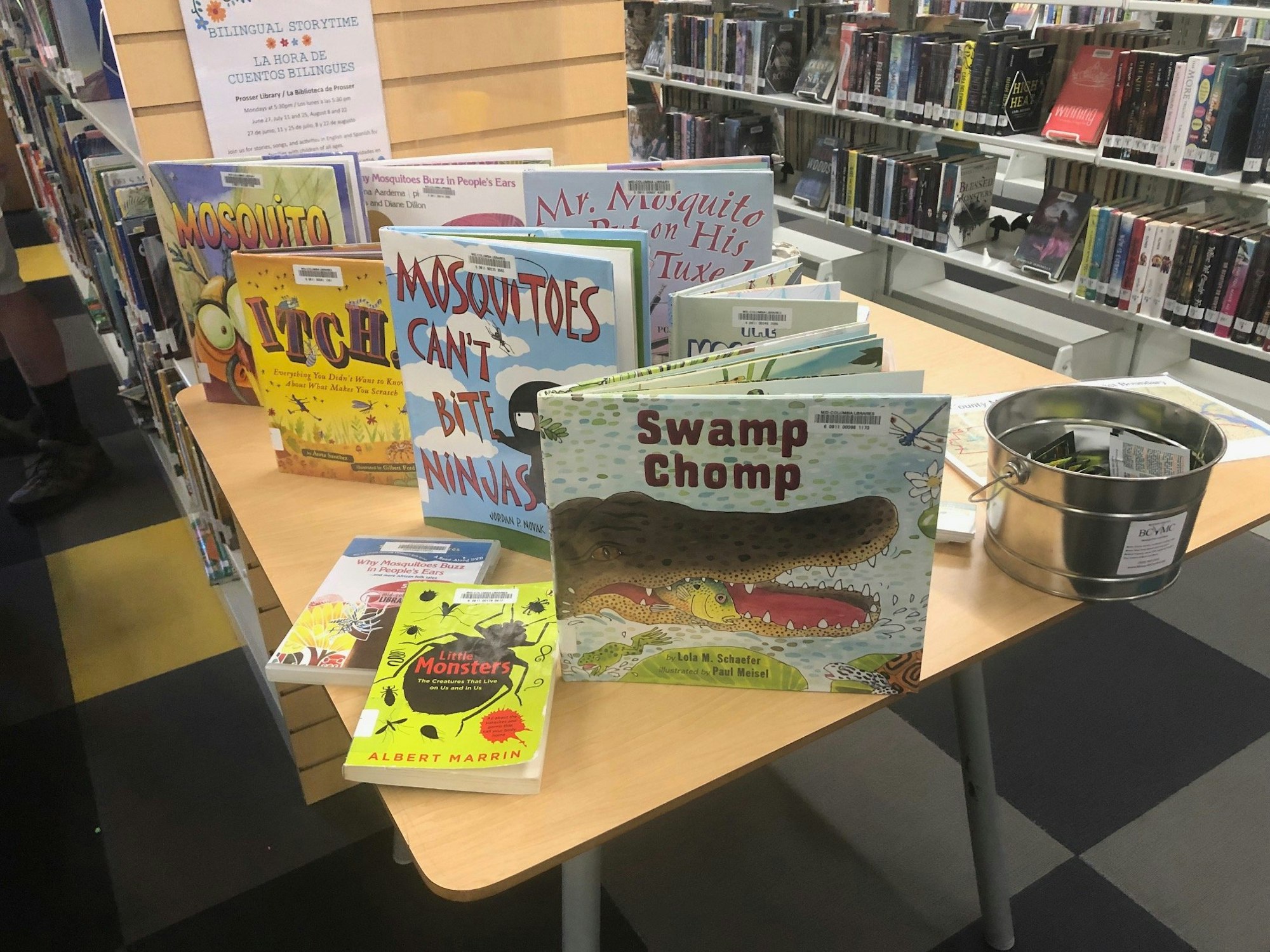 A collection of children's books displayed on a table in a library, some with mosquito themes, next to a metal bucket.