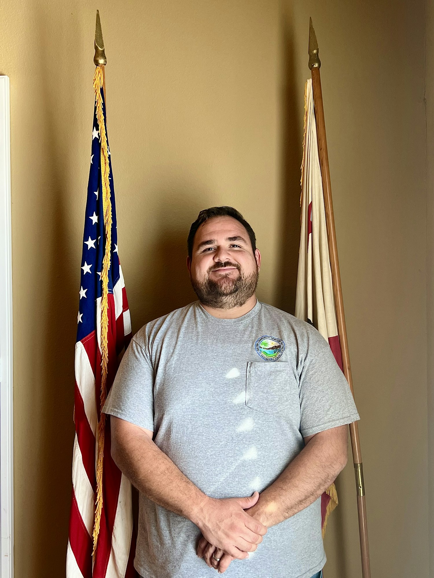 A man stands between an American flag and a California flag, smiling in a casual gray shirt against a neutral background.