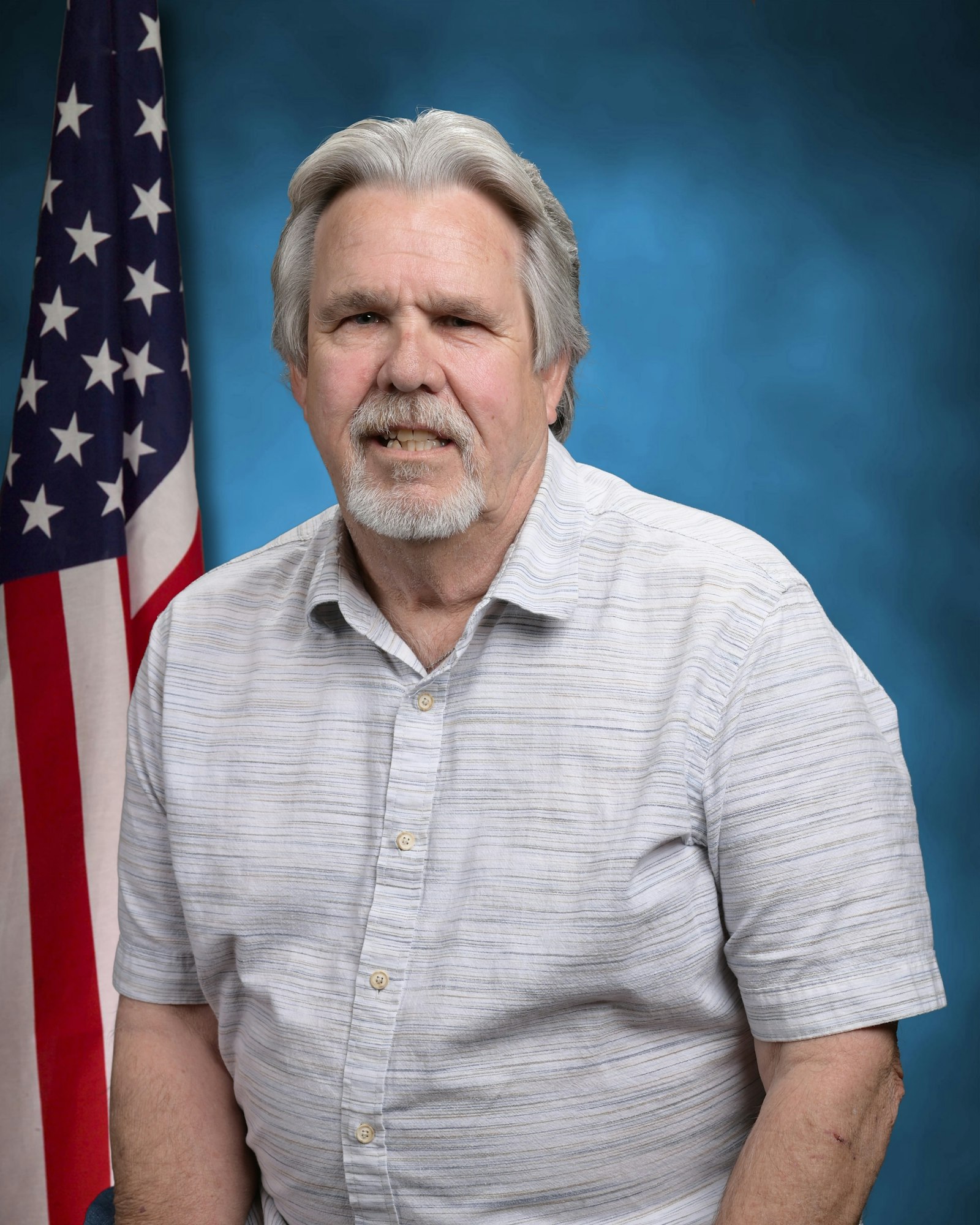 A man with gray hair and a gray beard wearing a light striped shirt poses in front of an American flag and blue background.