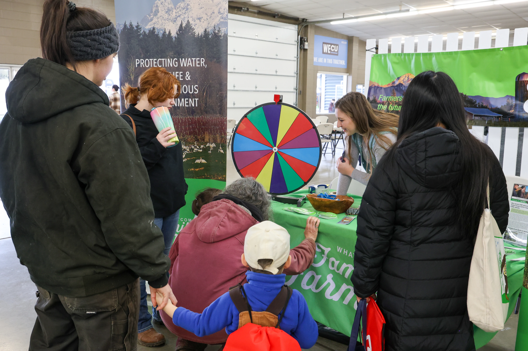 People gathered around a colorful prize wheel at an indoor event with educational banners.