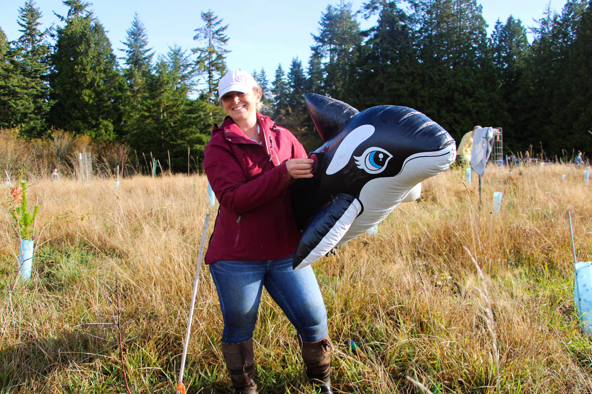 A woman in a maroon jacket holds an inflatable orca in a grassy field with trees in the background.