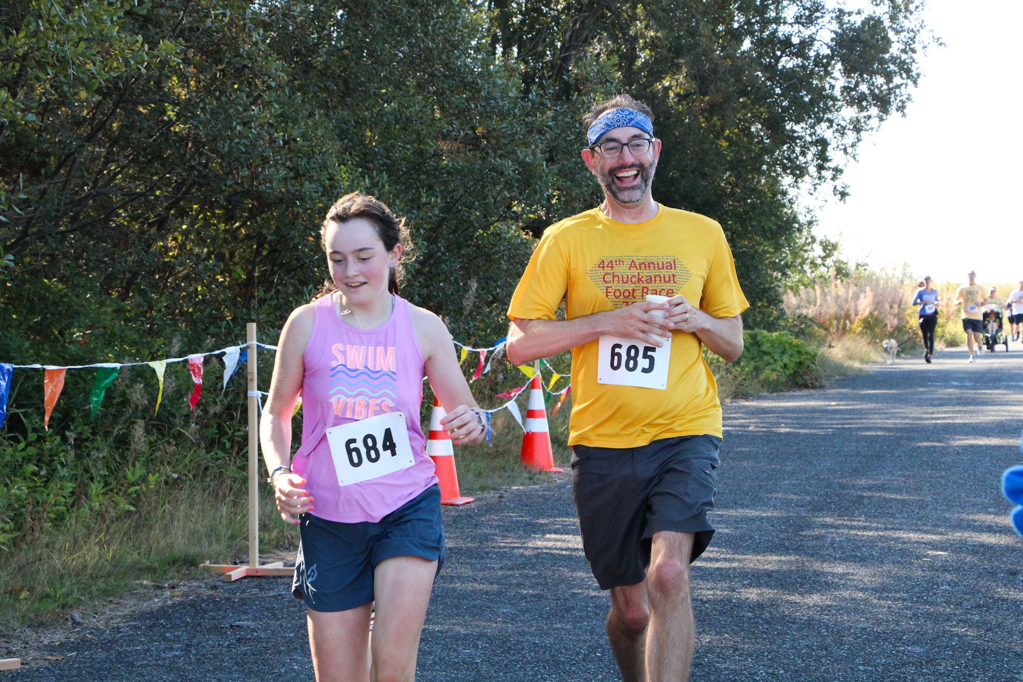 Two people running in an outdoor race with numbered bibs, surrounded by trees and colorful flags.