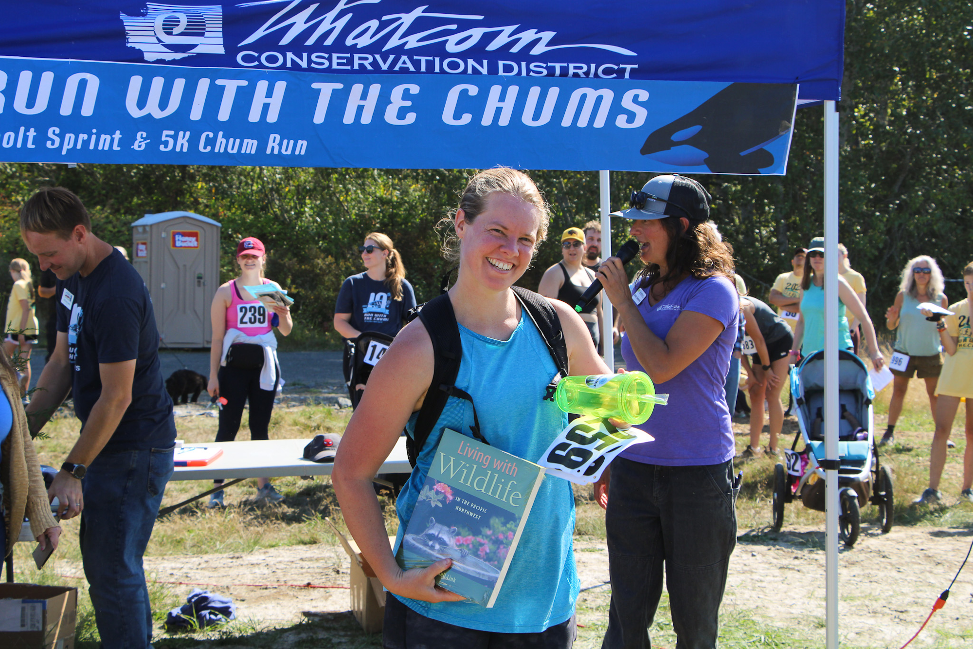 A smiling woman holding a book and water bottle at an outdoor event under a "Run with the Chums" banner.