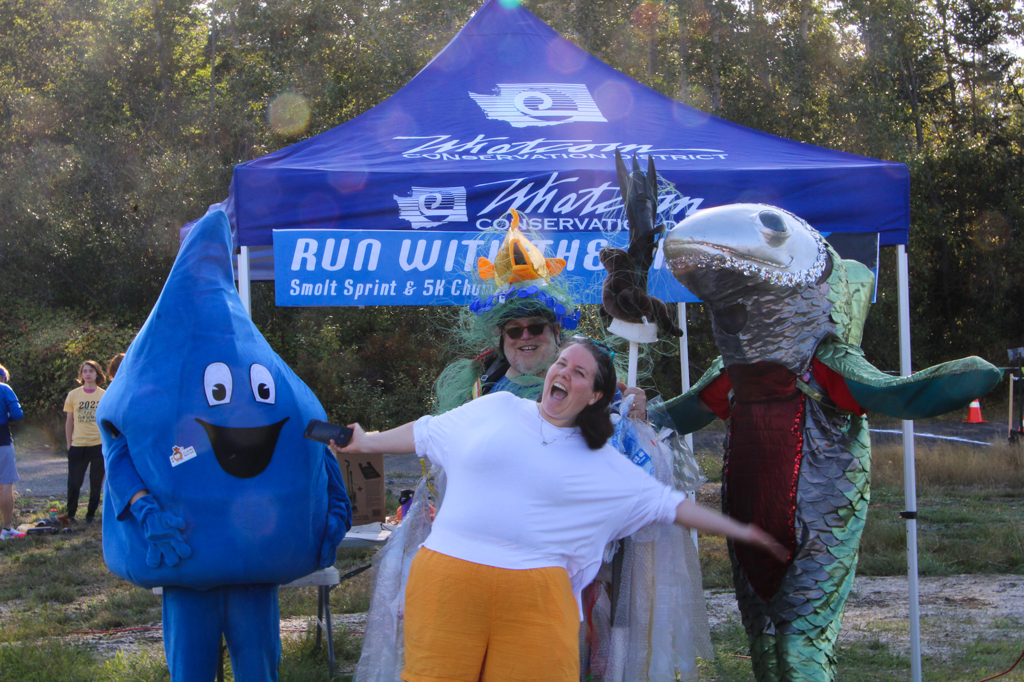 People in fun costumes pose under a "Run with the Fish" event tent in a wooded area.