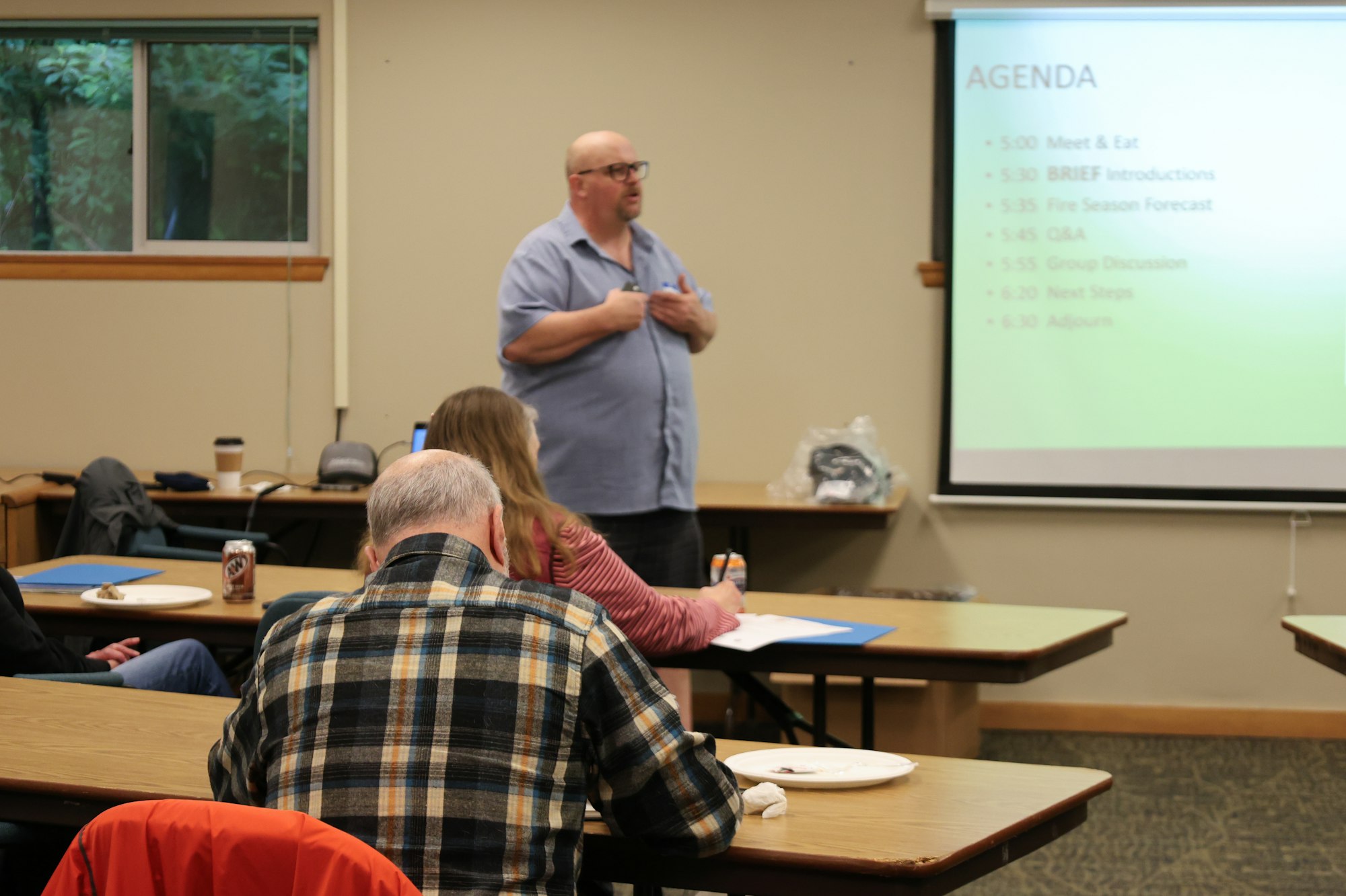 A presenter is discussing an agenda for a meeting about a fire season forecast with attendees seated at tables.