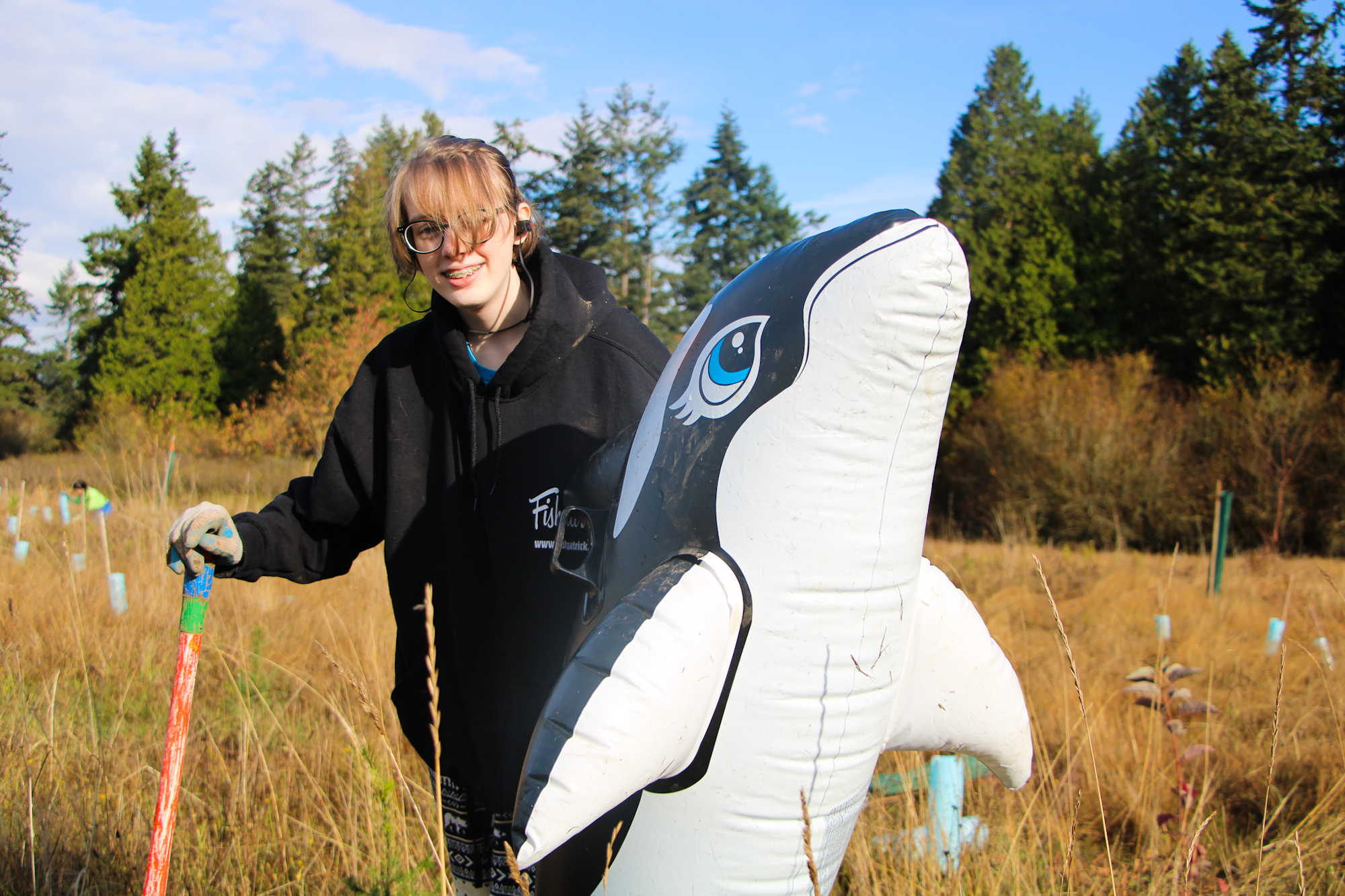 A person outdoors with a large inflatable orca and a garden tool, surrounded by tall grass and trees.