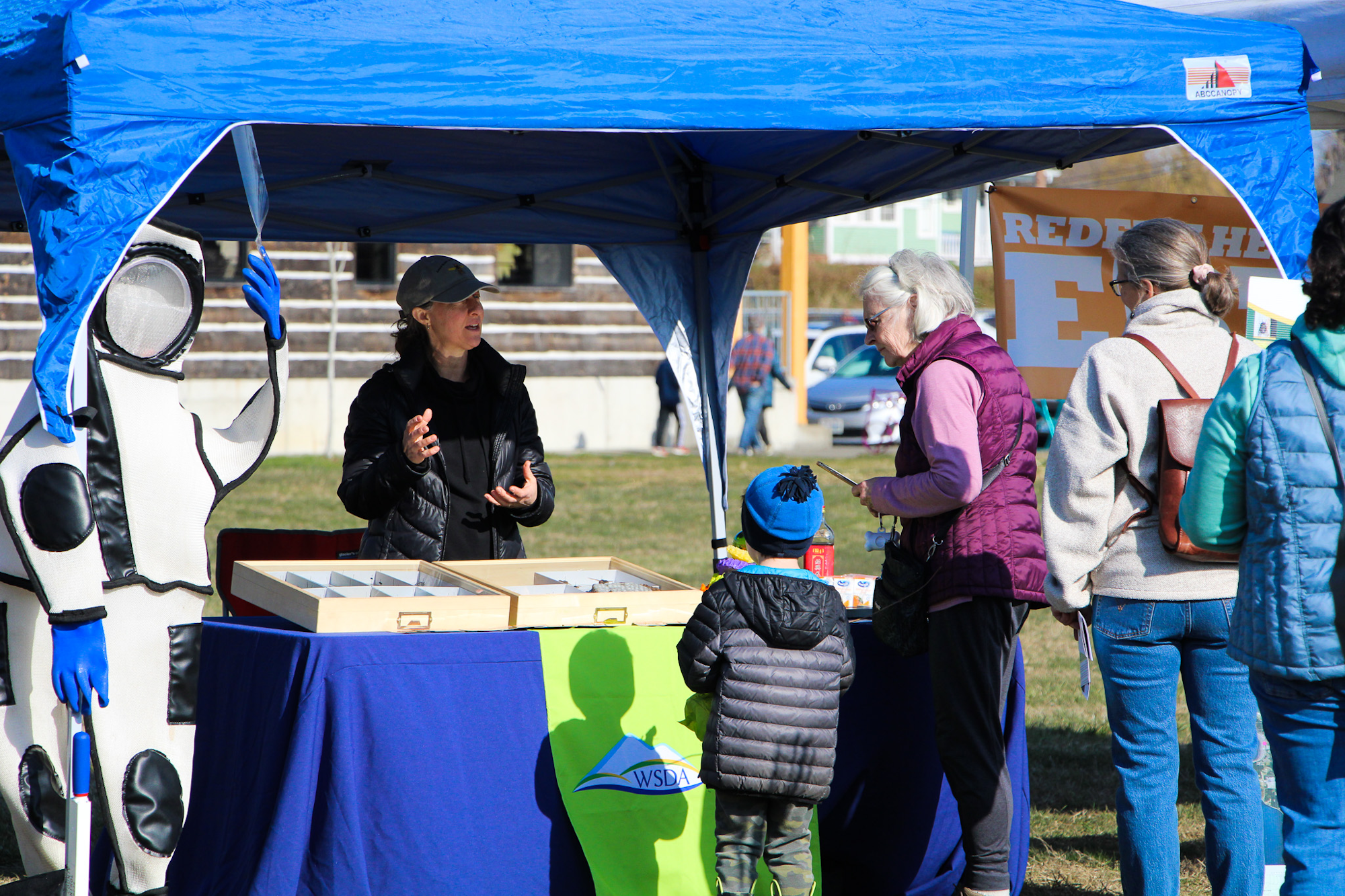 A person in a bee suit and another in a cap at an outdoor event booth, interacting with attendees under a blue canopy.