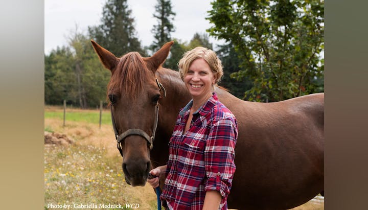 A person in a plaid shirt smiles next to a brown horse in an outdoor setting.