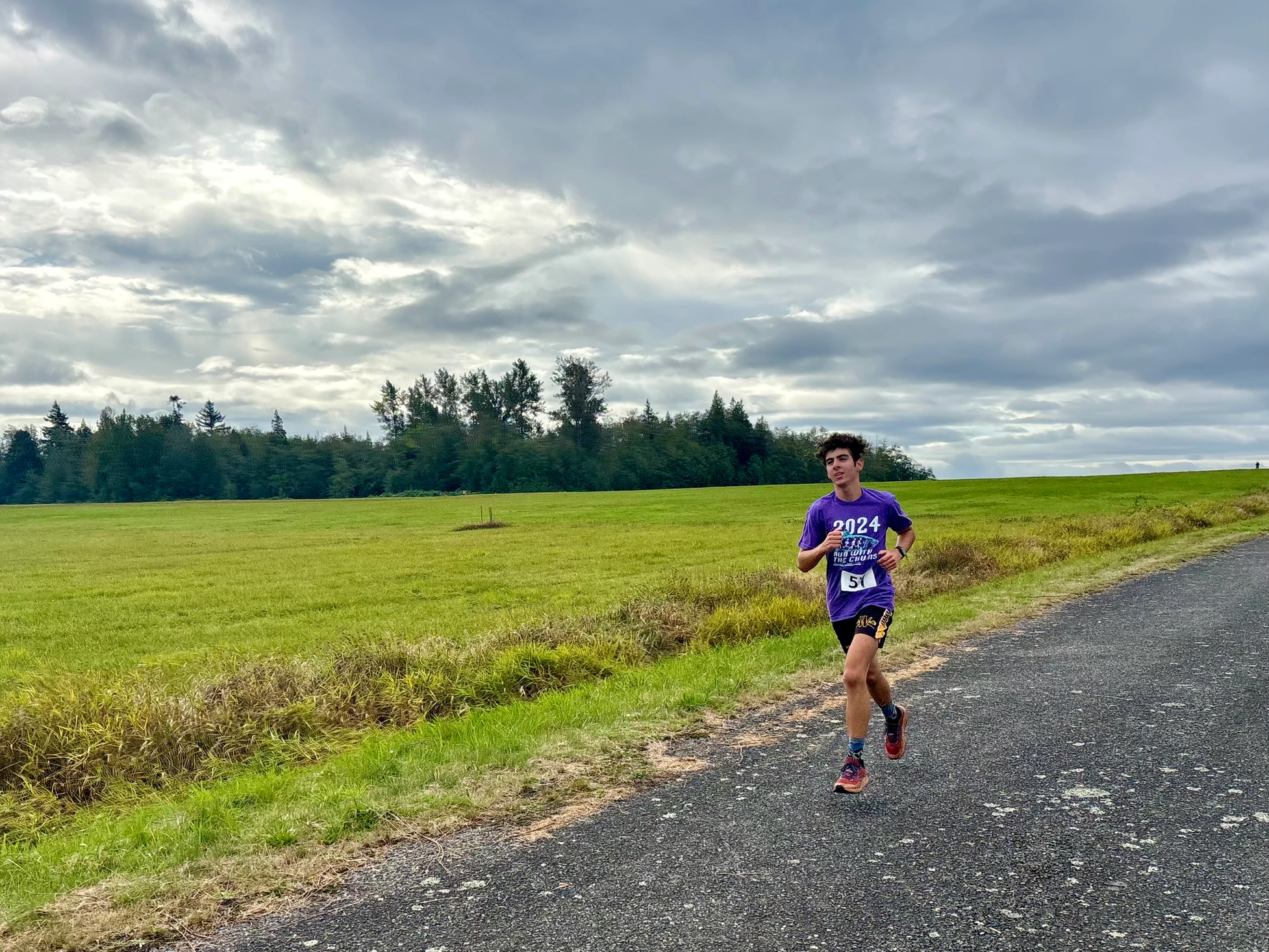 A person jogging along a rural road with green fields and cloudy skies in the background.