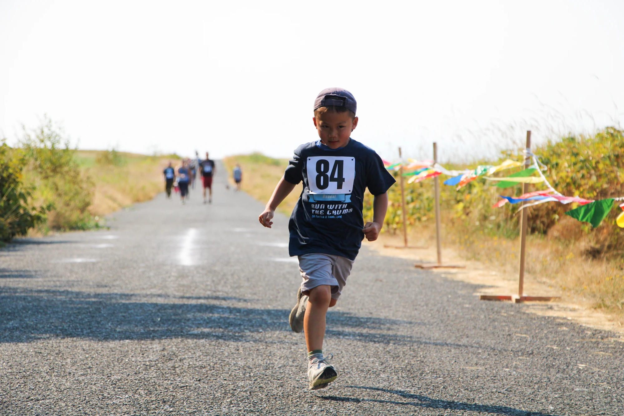 A young boy running on a path with a race bib number 84, other people in the distance, and colorful flags on the side.