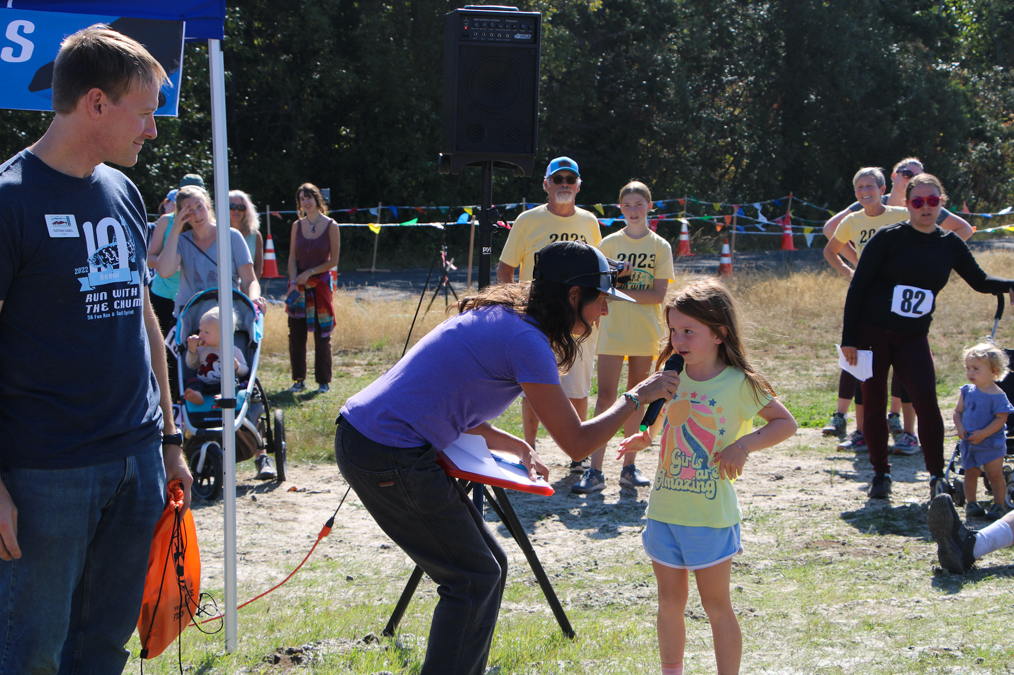 A person interviews a young girl outdoors; others watch nearby at a sports event.