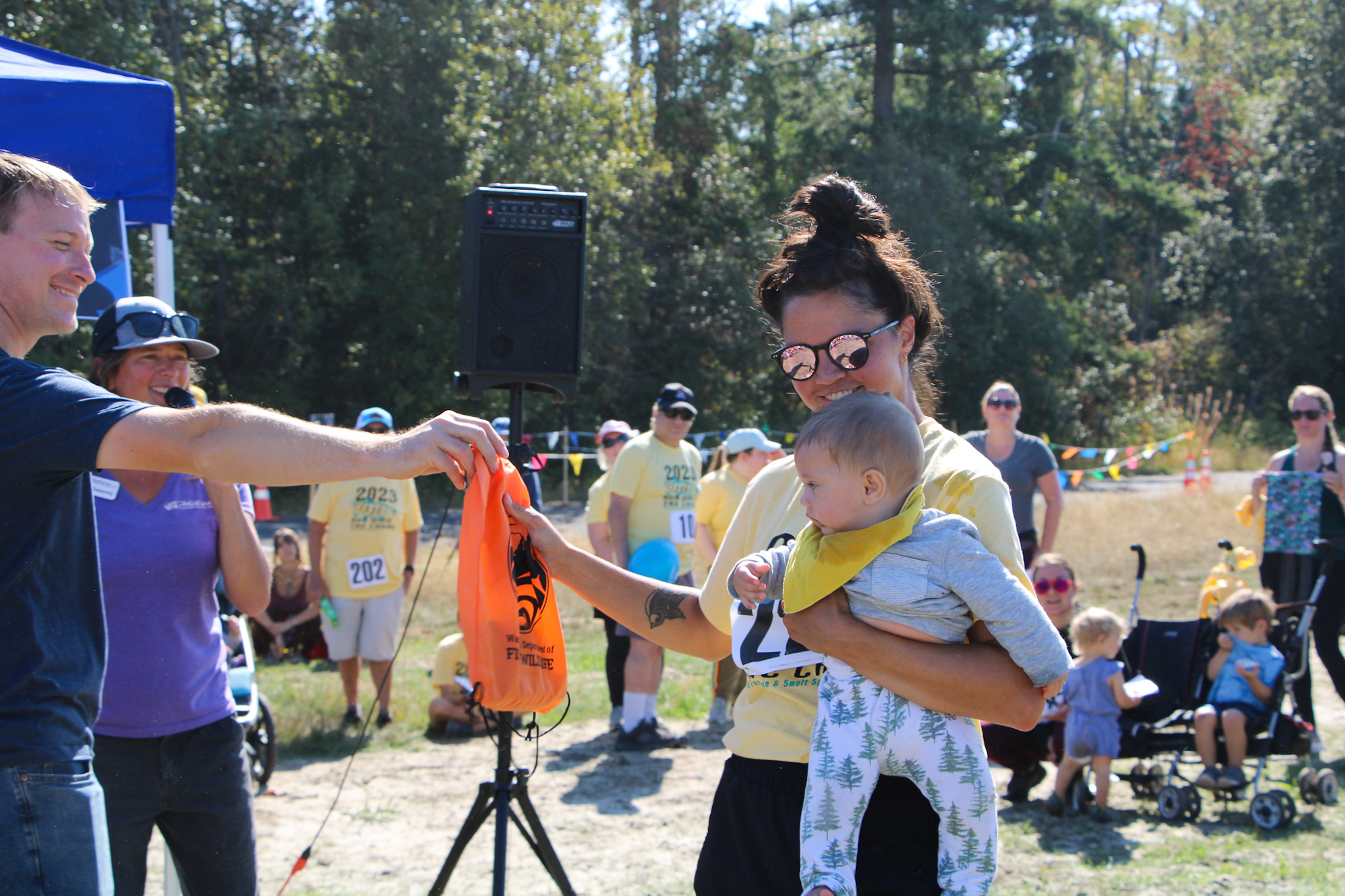 A woman holding a baby receives an orange bag from a man during an outdoor event, with onlookers in the background.