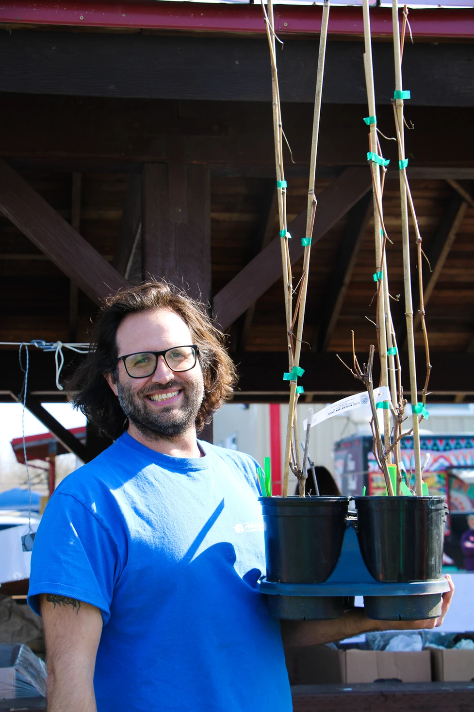 A person in glasses and a blue shirt smiling, holding potted plants with tall stakes.