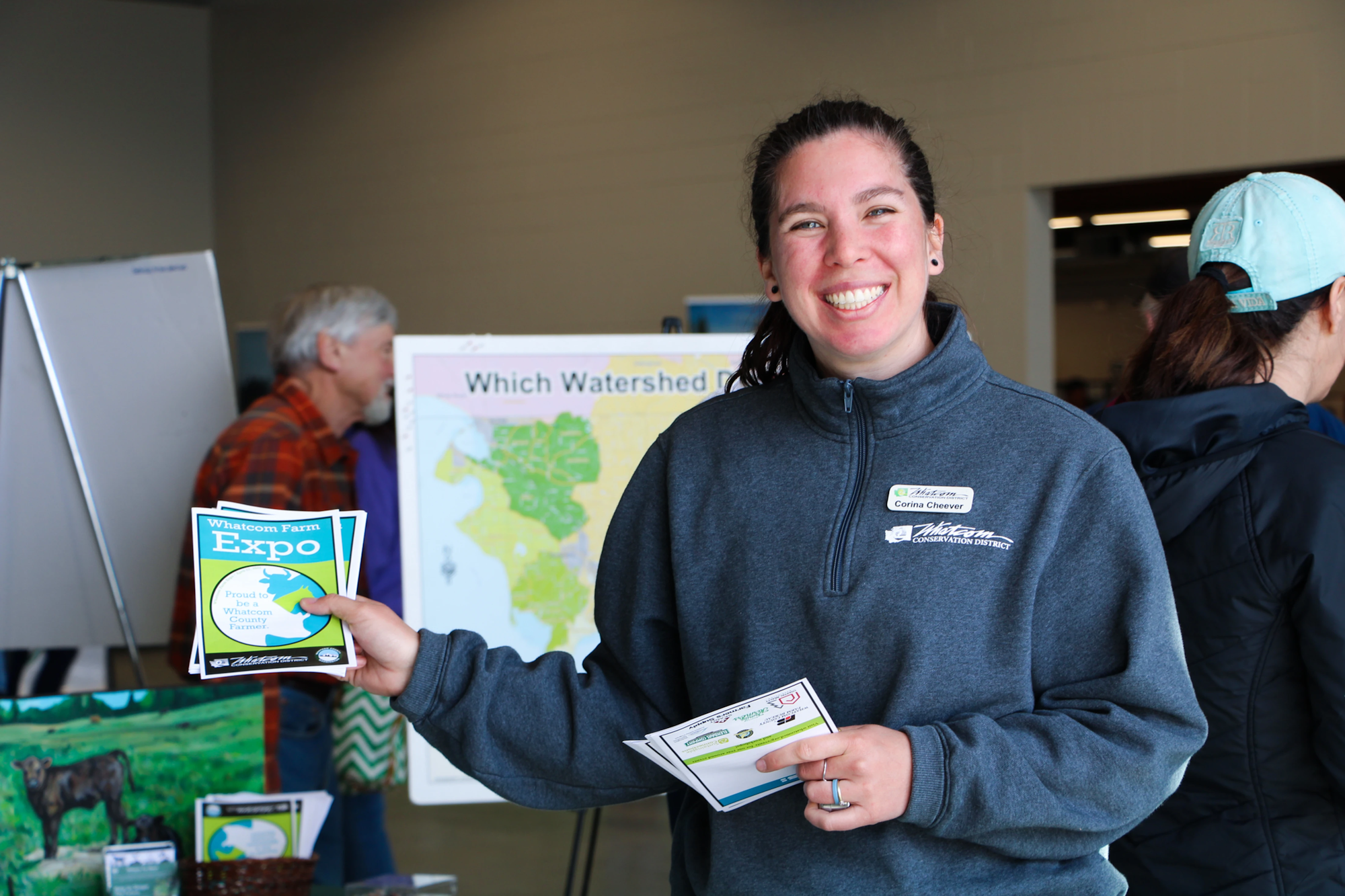 A person smiles while holding flyers for the Whatcom Farm Expo, with maps and people in the background.