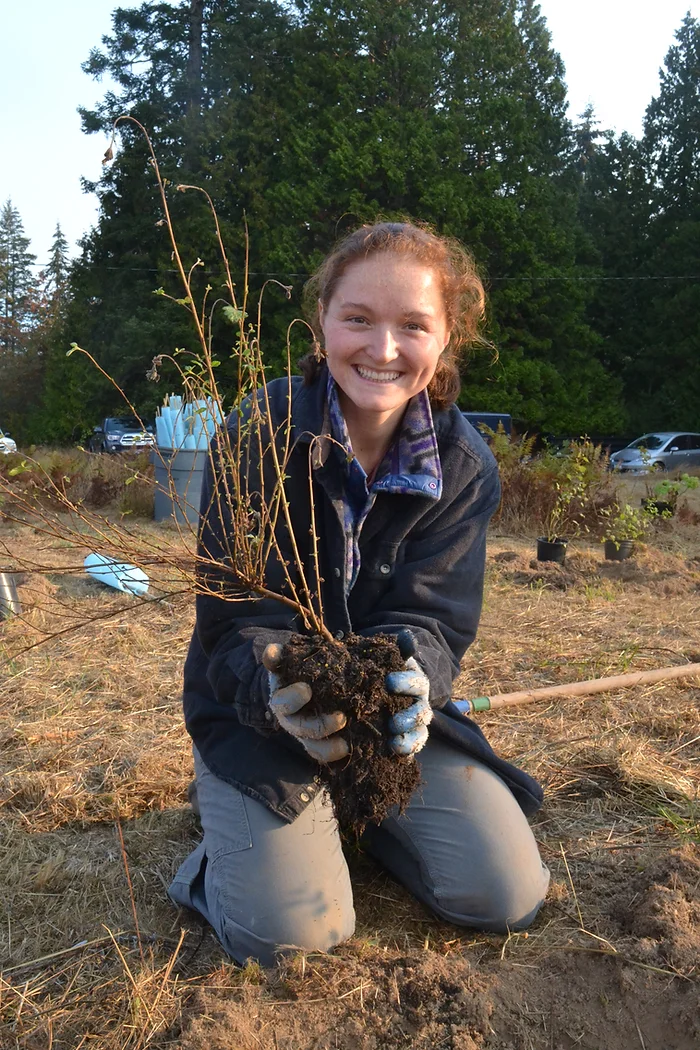 A person kneeling on grass, holding a small plant with soil in gloved hands, smiling in an outdoor setting.