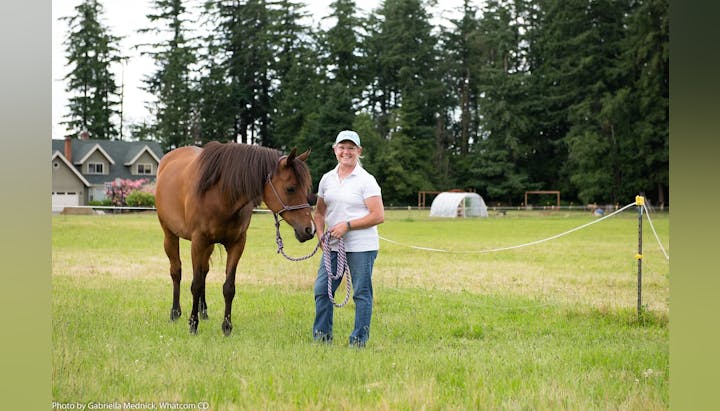 A person in a white shirt and cap stands smiling next to a brown horse in a grassy field, with trees and houses in the background.