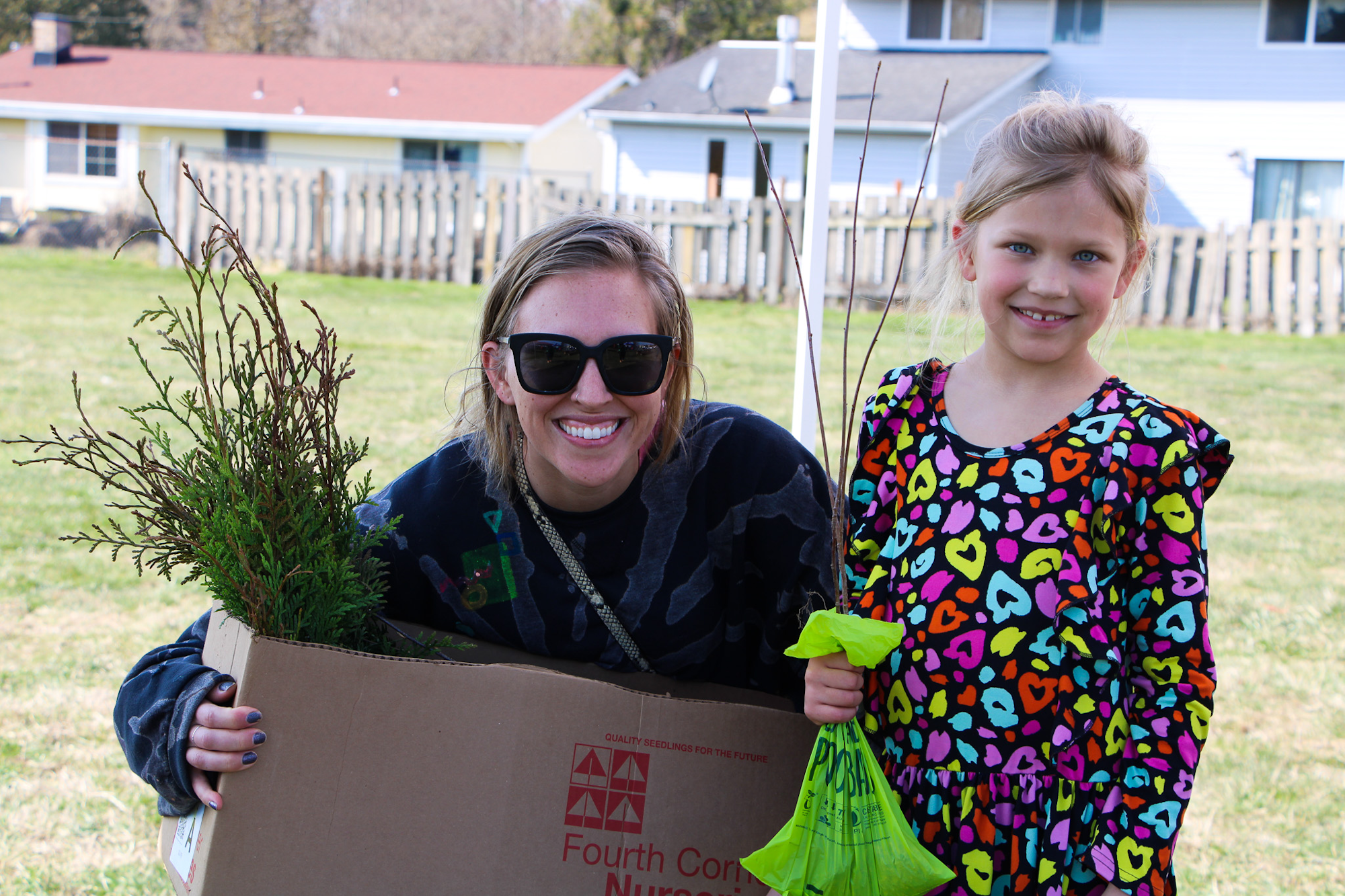 Two people smiling outdoors; one holds a box with plants, the other holds a green bag.