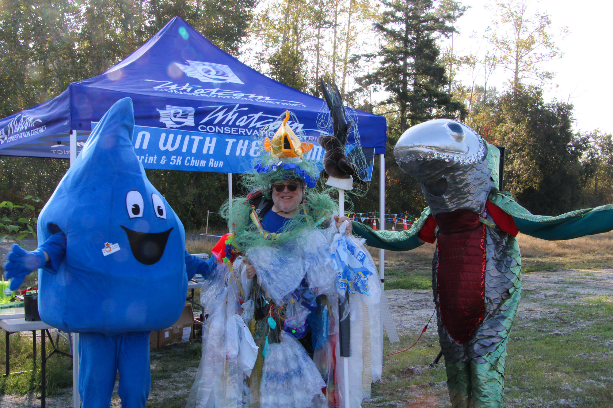 Three costumed mascots—a water droplet, a fish, and a person dressed in recycled materials—at a conservation event.