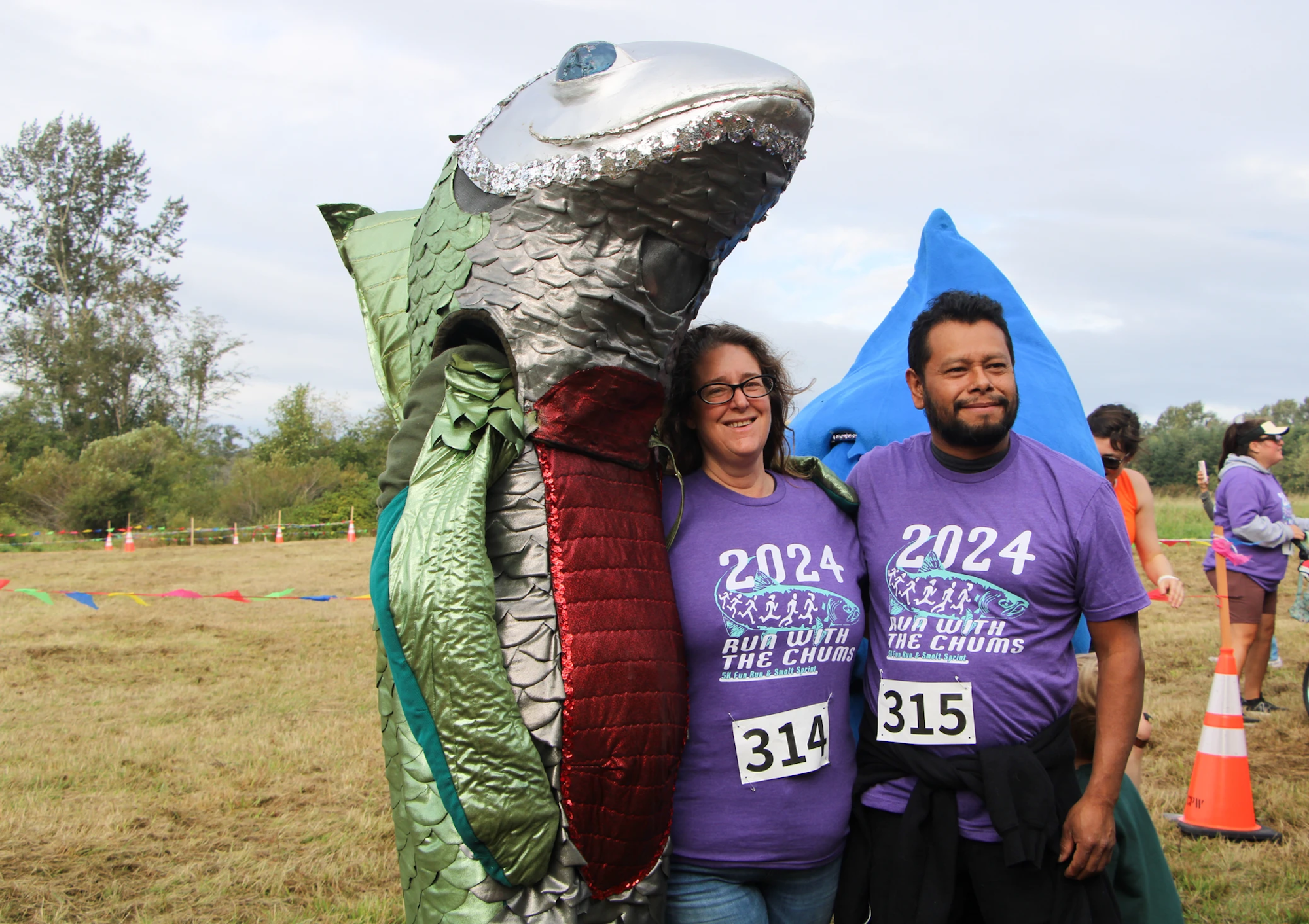 People in fish costumes and purple "Run with the Chums 2024" shirts posing outdoors at an event.
