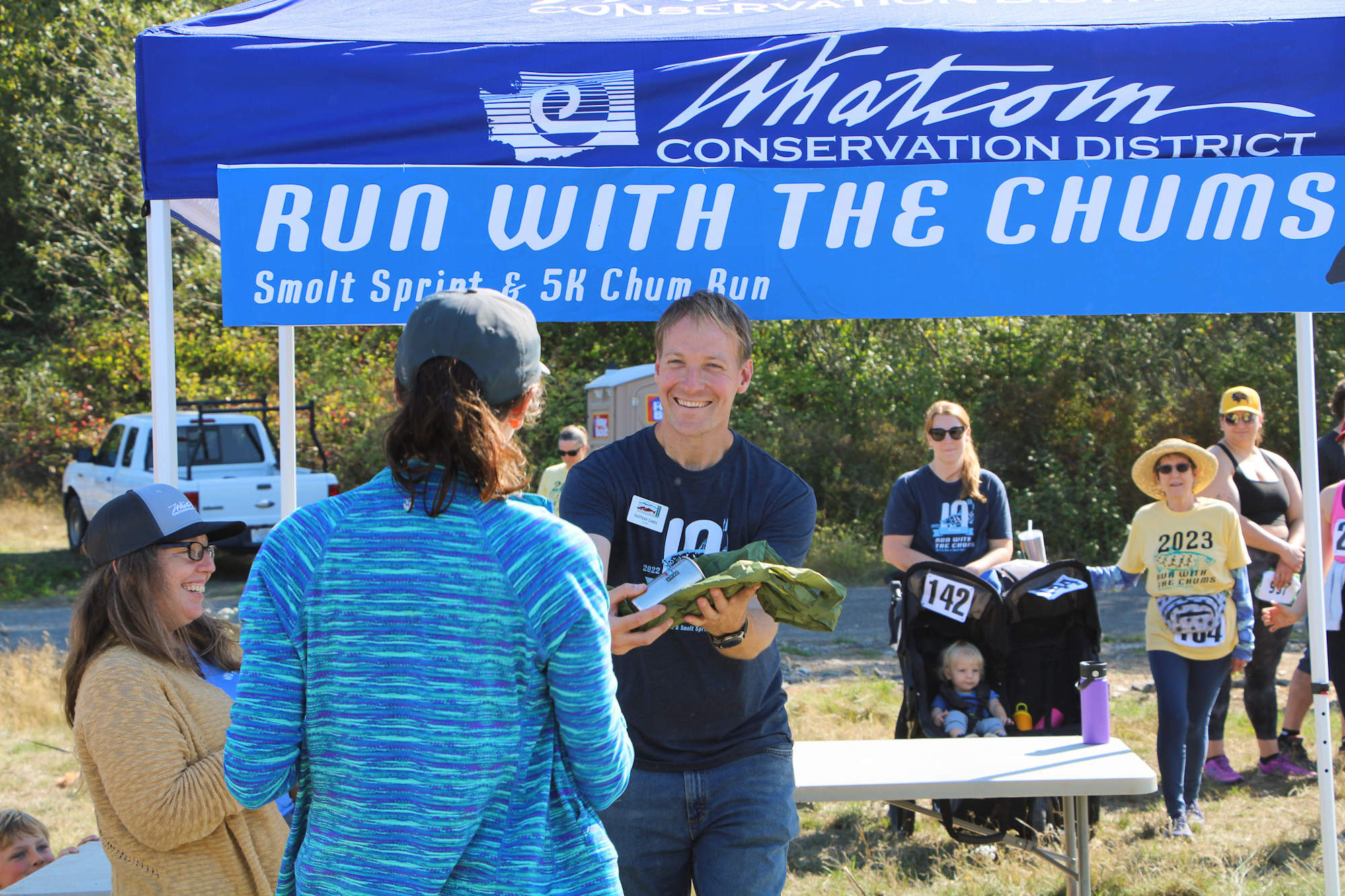 Outdoor event with people under a tent labeled "Run with the Chums," featuring a race and awards.