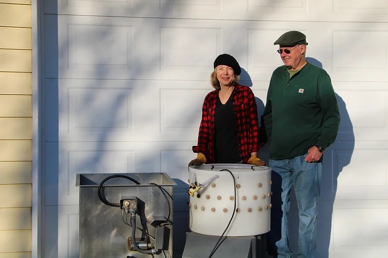 Two people stand outdoors near a metal drum and equipment in front of a garage door.