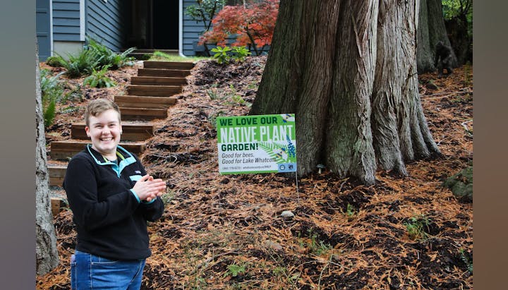 Person smiling near a tree with a sign: "We love our native plant garden! Good for bees. Good for Lake Whatcom."