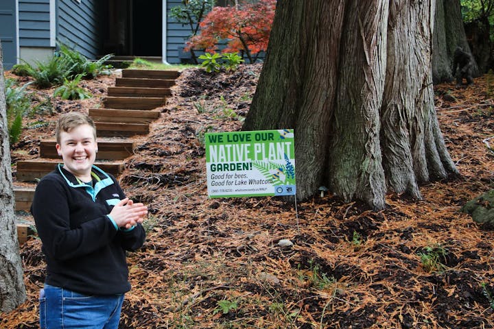 Person smiling near a tree with a sign: "We love our native plant garden! Good for bees. Good for Lake Whatcom."