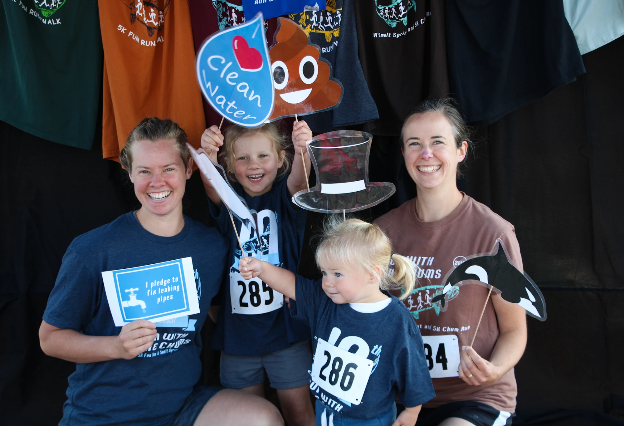 A family at a fun run event posing with signs about clean water and numbers, smiling in front of a backdrop of t-shirts.