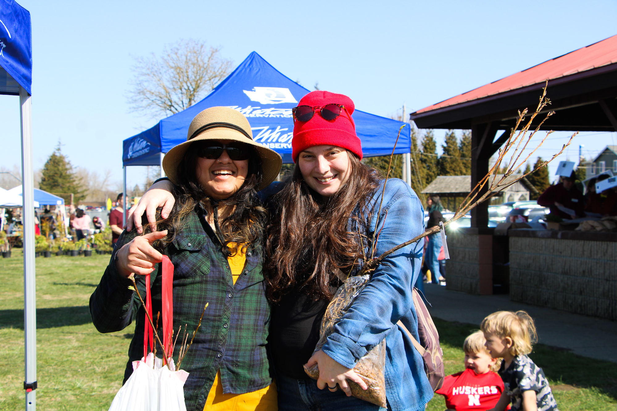 Two people smiling at an outdoor market, one holding a bag with twigs.