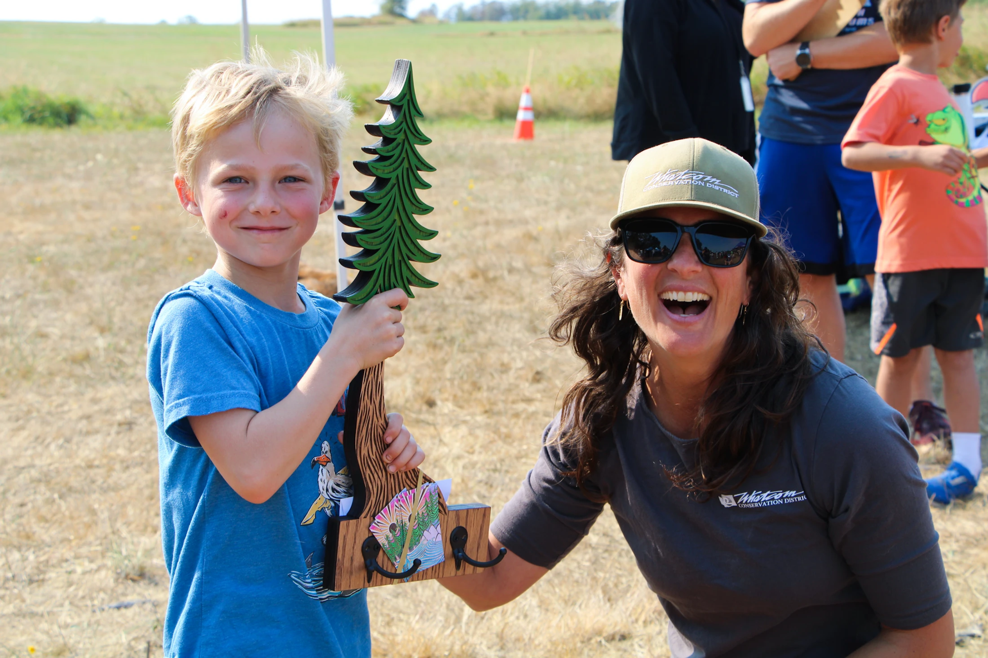 A smiling child holding a tree-shaped object with a woman in a cap and sunglasses posing next to him outdoors.