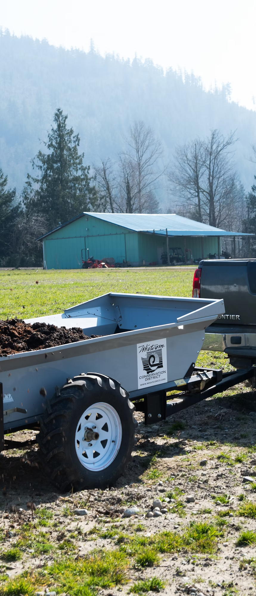 A trailer attached to a vehicle, filled with soil, on a farm beside a barn and trees.