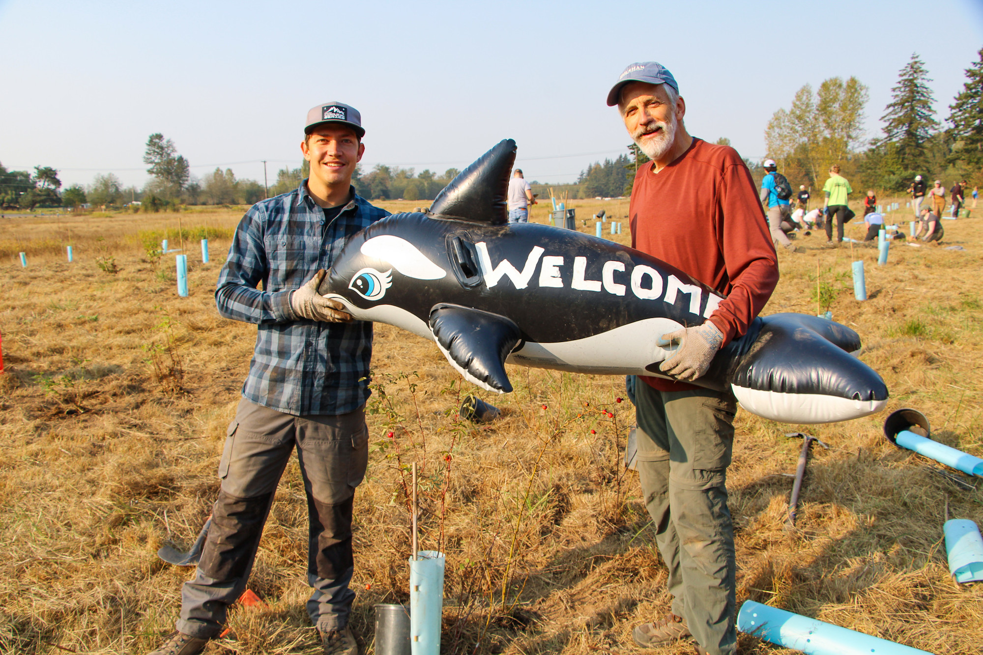 Two people holding an inflatable orca with "Welcome" on its side in a field, others planting in the background.