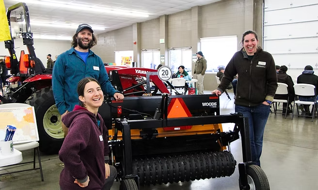 Three people smiling next to farming equipment at an indoor event with tractors and seated attendees.