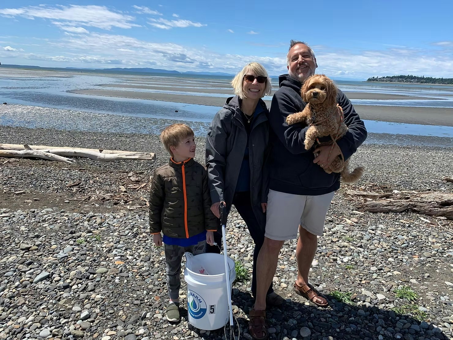 Three people and a dog on a rocky beach, holding a bucket with a scenic coast in the background.